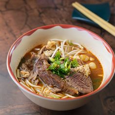 a bowl filled with noodles and meat on top of a wooden table next to chopsticks