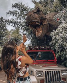 a girl reaching for a dinosaur statue on top of a jeep in front of a tree