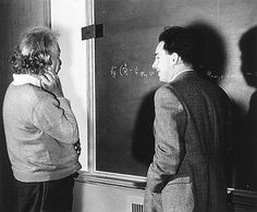 two men standing in front of a blackboard with writing on it and one man wearing a coat