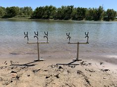 two metal sculptures sitting on top of a sandy beach next to water with trees in the background