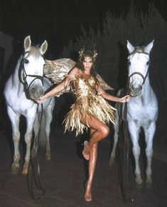 a woman in a gold dress standing next to two white and brown horses at night