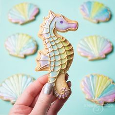 a hand holding a seahorse cookie in front of some seashells on a blue background