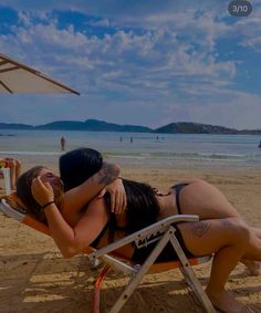 two women in bikinis are sitting on a beach chair with an umbrella over them