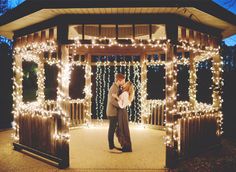 a man and woman standing in front of a gazebo covered with christmas lights