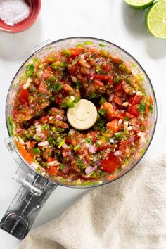 a food processor filled with chopped vegetables on top of a white table next to limes