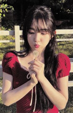 a young woman blowing on a dandelion in front of a white picket fence