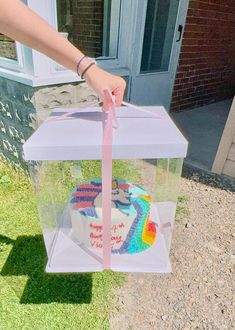 a person holding a cake in a clear box with pink ribbon on the outside of it