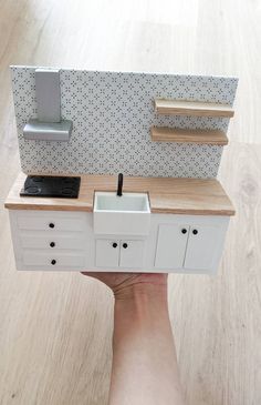 a hand holding a miniature model of a kitchen with sink and countertop, in the shape of a doll house