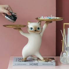 a white cat figurine holding a plate on top of a table next to a pink wall