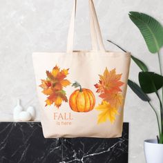 a canvas bag with autumn leaves and pumpkins painted on it, sitting next to a potted plant