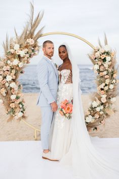 a bride and groom standing under an arch on the beach with their bouquets in front of them