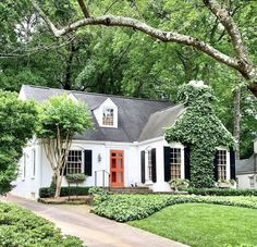 a white house with black shutters and red door surrounded by greenery in the front yard