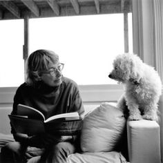 a person sitting on a couch reading a book with a dog standing next to them