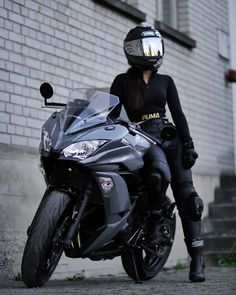 a woman sitting on top of a black motorcycle next to a brick building and wearing a helmet