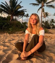 a woman sitting on top of a sandy beach next to palm tree's at sunset