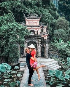 a man and woman standing on a bridge in front of water lilies, surrounded by greenery