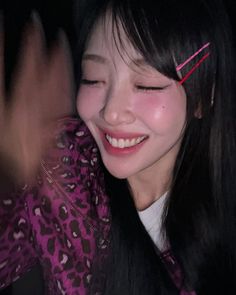 a woman with long black hair and pink shirt smiling at the camera while holding her hand up to her face