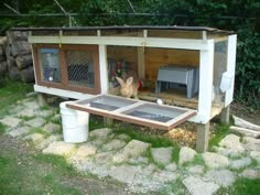 a small chicken coop in the middle of some rocks and grass with a cat sitting on it's side