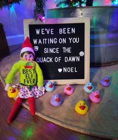 an elf is sitting next to a sign that says, we've been waiting on you since the quack of dawn noel