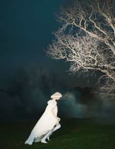 a woman standing in front of a tree at night wearing a white dress and hat