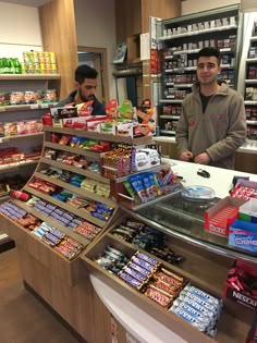 two men standing in front of a store counter filled with snacks and candy bar items