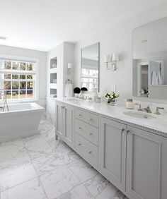 a white bathroom with marble flooring and large tub in the center, surrounded by two windows