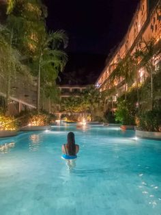 a woman sitting in the middle of a swimming pool at night with buildings behind her