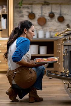 a woman kneeling down in front of an oven holding a tray with food on it