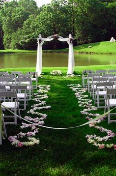 an outdoor ceremony setup with white chairs and pink flowers on the grass, near a pond