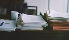 stacks of papers on a desk next to a coffee cup and pen with a window in the background