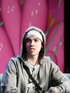 a young man sitting in front of a pink wall wearing headphones and a hoodie