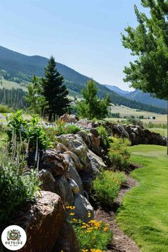 a stone wall in the middle of a grassy area with flowers and trees on either side