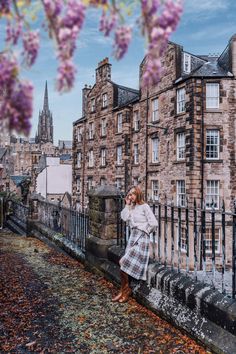 a woman standing on the side of a bridge in front of some buildings with purple flowers