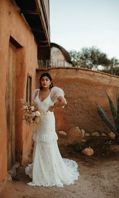 a woman standing in front of a building with a bouquet on her hand and wearing a white dress