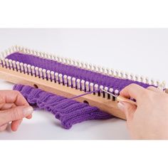 two hands are working on an object with purple yarn and wooden dowels, while another hand is holding a pair of scissors