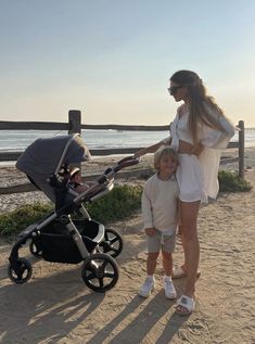 a woman standing next to a baby in a stroller on the beach with her son