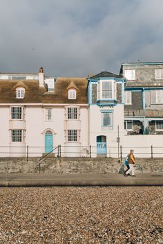 two people walking on the beach in front of some houses with blue doors and windows