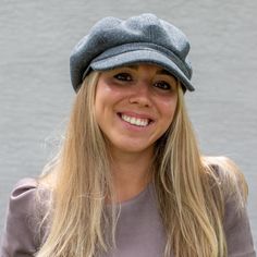 a woman with long blonde hair wearing a gray hat and smiling at the camera while standing in front of a wall