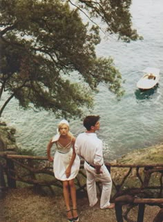 a man and woman standing next to each other on a hill overlooking the ocean with boats in the water