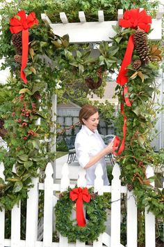 a woman standing in front of a white picket fence with wreaths and red bows