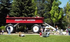 people are sitting on the grass in front of a large red truck with white wheels