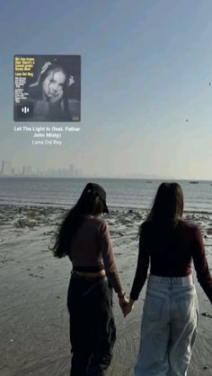 two women walking on the beach holding hands and looking out at the ocean with an ad in the background