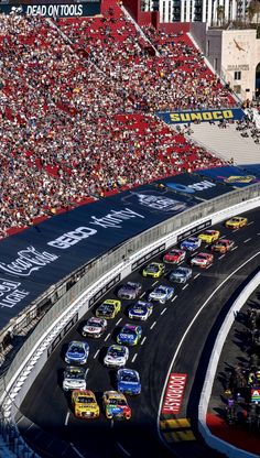 a group of cars driving down a race track in front of a large crowd at a sporting event