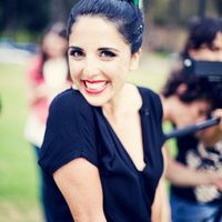 a woman is smiling and holding a camera behind her back to the camera, with other people in the background