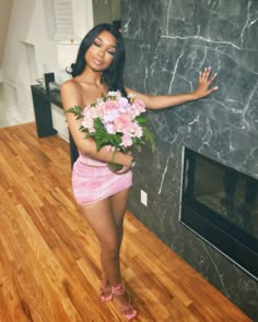 a woman in pink dress holding flowers standing next to a blackboard with marble wall and fireplace