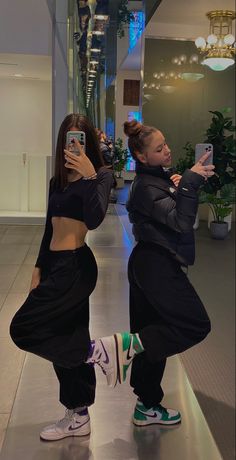 two young women taking pictures with their cell phones in an office building hallway, both wearing black clothing and green sneakers