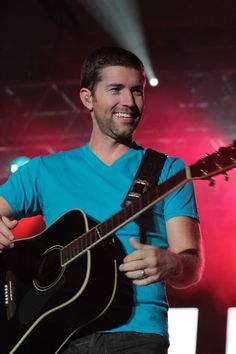 a man playing an acoustic guitar on stage at a music festival or concert with lights in the background