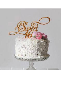 a white cake with gold lettering and pink flowers on top is sitting on a glass stand