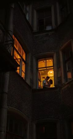 two people standing in the window of an apartment building at night with their arms around each other