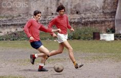 two young men playing soccer on a dirt field